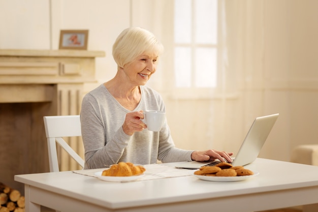 Warm talk. Delighted pensioner female keeping smile on her face and holding cup in right hand while typing message