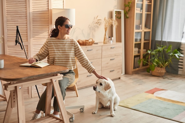 Теплый тонированный портрет в полный рост современной слепой женщины, сидящей за столом в уютном домашнем интерьере и ласкающей собаку-поводыря, копировальное пространство