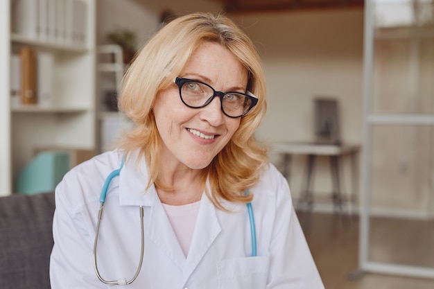 Warm-toned head and shoulders portrait of mature female smiling at camera while working in modern clinic, copy space