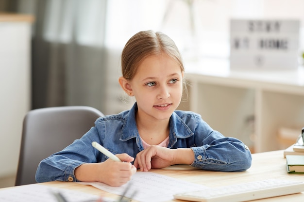 Ritratto dai toni caldi della bambina sveglia che sorride mentre fa i compiti alla scrivania e studia a casa in interni accoglienti