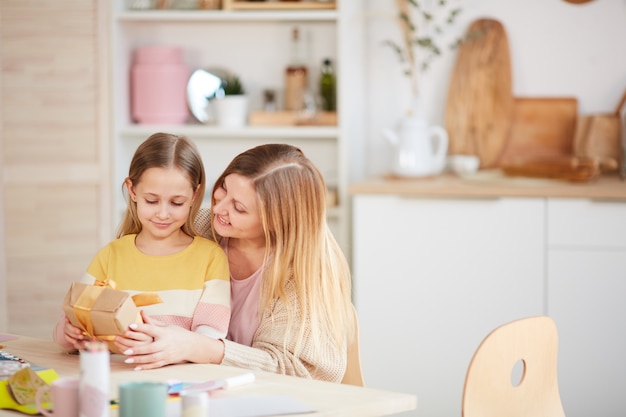 Warm-toned portrait of happy mother hugging daughter while opening presents at kitchen table, copy space