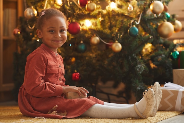 Warm toned portrait of smiling African-American girl opening Christmas presents while sitting by tree at home, copy space