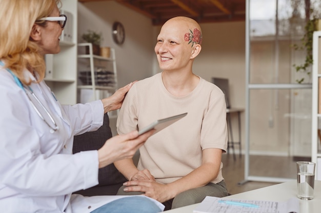 Ritratto dai toni caldi della donna calva sorridente che parla alla dottoressa durante la consultazione su alopecia e recupero del cancro, copia dello spazio