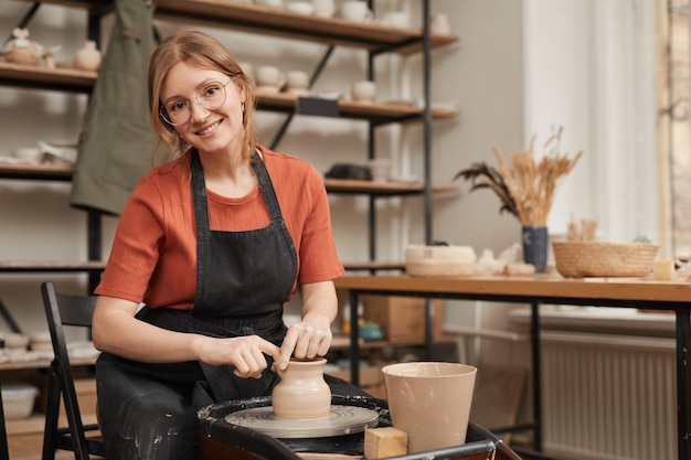 Warm toned portrait of young female potter smiling at camera while working on pottery wheel in workshop and enjoying arts and crafts, copy space