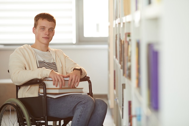 Dai toni caldi ritratto di giovane uomo con sedia a rotelle a scuola mentre in biblioteca illuminata dalla luce solare