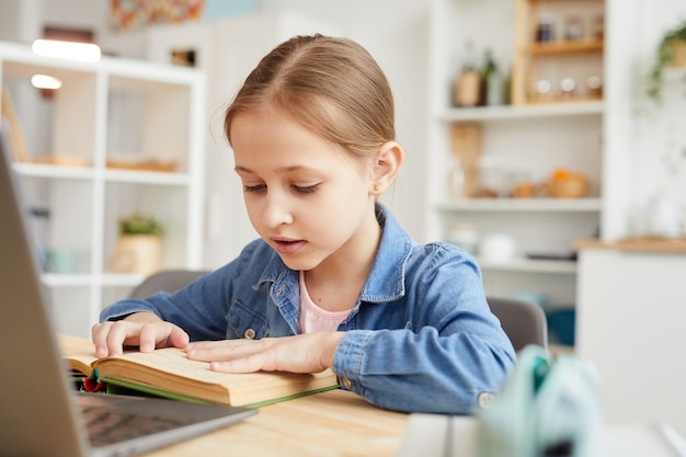 Warm-toned portret van schattig klein meisje tekst lezen op laptop camera tijdens het studeren online thuis in gezellig interieur, kopieer ruimte