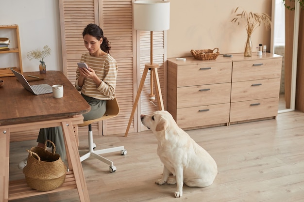 Warm toned wide angle portrait of modern young woman using smartphone at home office with dog sitting by her in cozy interior, copy space