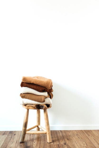 Warm winter women woolen knitted sweaters, pullovers stack on wooden stool at white. Female modern clothes fashion blog concept.