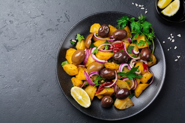 Warme aardappelsalade met olijven, peper, peterselie en rode ui op zwarte ceramische plaat op donkere concrete oppervlakte