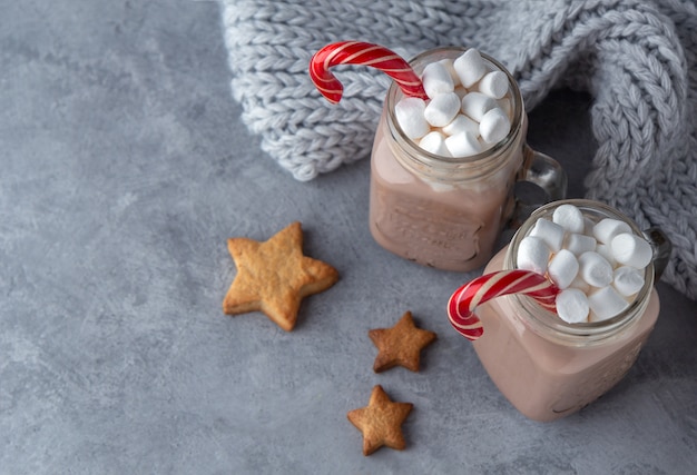 Warme chocolademelk met marshmallows en een suikerriet in glazen mokken op een grijze achtergrond met een gebreide sjaal.