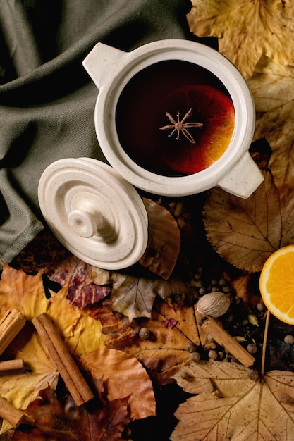 Warme glühwein in keramische pot met kruiden, sinaasappel- en herfstbladeren over donker linnen tafelkleed. Plat leggen. Gezellige warme alcoholische dranken. plat leggen