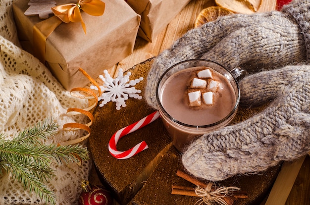 Warme kerstdrank van cacao met marshmallows. Handen in wanten houden een mok met een drankje op tafel met geschenken en kerstboomtakken.