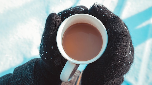 Warme kop warme koffie opwarming van de aarde in de handen in wanten. womens handen op de achtergrond van sneeuw, winter