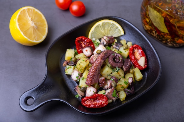 Warme salade met octopus, aardappelen, zongedroogde tomaten, kappertjes en citroen in een zwart pannetje. Close-up, zwarte achtergrond.