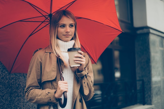 ホットコーヒーでウォーミングアップ。通りに立っている間傘とコーヒーカップを運ぶ魅力的な若い女性