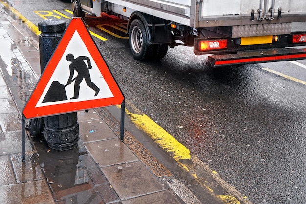 Warning roadworks sign at wet road next to pavement, van moving
in background over bus stop writing on road