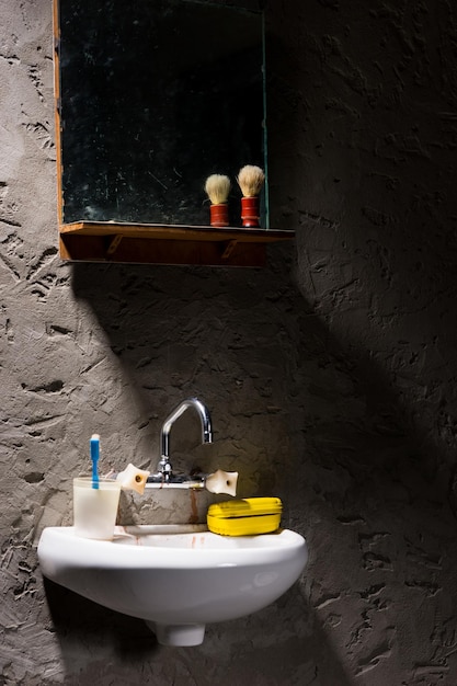 Washbasin with a glass for a toothbrush and a soap box on it under the mirror with shave brush on the shelf in a prison cell