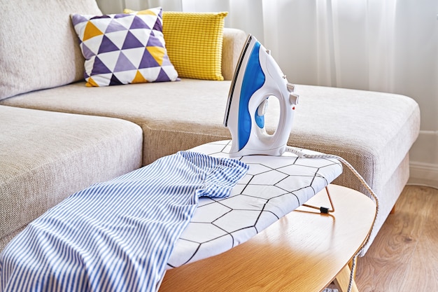 Washed blue shirt on tabletop ironing board and iron in small living room with sofa, household routine concept