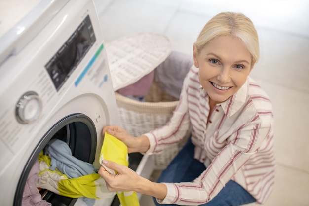 Washing. Blonde woman putting clothes into washing machine