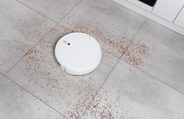 Washing White robot vacuum cleaner device cleaning floor from crumbs groats at home on utonite tiles in the kitchen