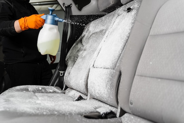 Wasmiddel op textielstoelen in auto-interieur sproeien voor chemisch reinigen Professionele reiniger in handschoenen giet wasmiddel op stoel in auto voor het verwijderen van vlekken en vuil