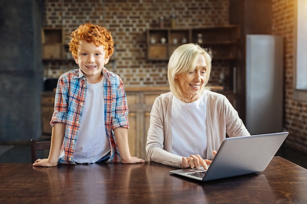 Wat doe je hier. Schattige roodharige jongen lachend in de camera terwijl leunend op een tafel naast zijn vrolijke oma met behulp van haar laptop thuis.