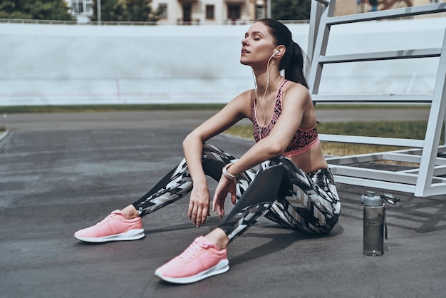 Wat een geweldig gevoel! Mooie jonge vrouw in sportkleding die een pauze neemt terwijl ze buiten zit