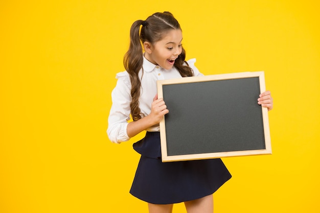 Wat is het Verrast meisje met schoolbord op gele achtergrond Klein kind schoolbord voorbereiden op les Klein kind met schoolbord Leeg bord voor schooladvertentie kopie ruimte