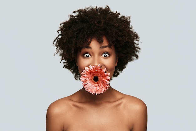 Wat? Verrast jonge Afrikaanse vrouw die naar de camera kijkt en bloem in de mond houdt terwijl ze tegen een grijze achtergrond staat