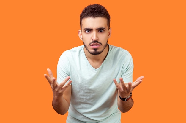 Wat wil je. Portret van ontevreden geërgerde brunette man met baard in casual wit t-shirt staande met opgeheven handen, gekke verontwaardigde uitdrukking. indoor studio-opname geïsoleerd op oranje achtergrond