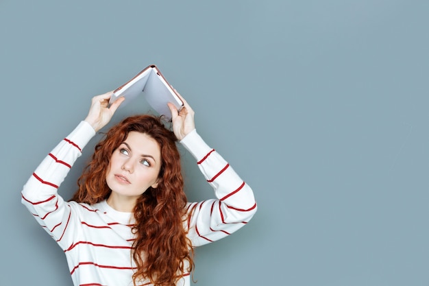 Wat zit erin. Leuke aangename jonge vrouw die een boek boven haar hoofd steekt en ernaar kijkt terwijl ze naar iets zoekt