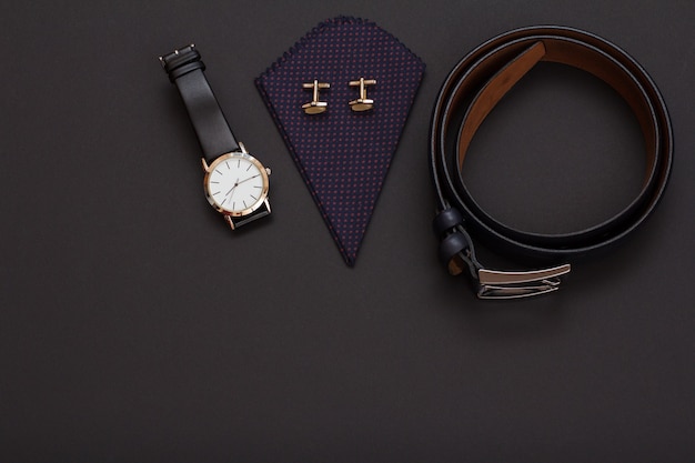 Watch with a black leather strap, handkerchief with cufflinks and leather belt with metal buckle on black background. Accessories for men. Top view.