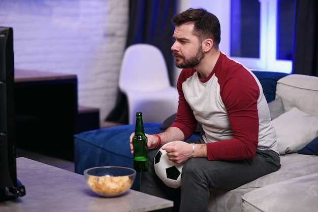 ゲームを見ています。テレビを見ながらビールを飲んだり、軽食を食べたりする陽気な若い男性