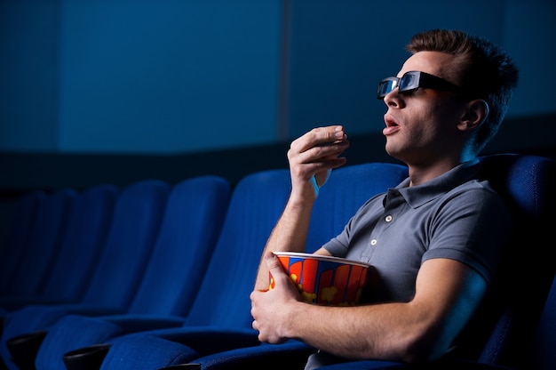 Watching three-dimensional movie. Excited young man in three-dimensional glasses eating popcorn and watching movie while sitting at the cinema