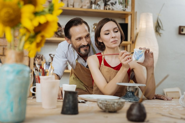 Watching wife. Smiling handsome bearded man watching his appealing cheerful wife decorating clay pot