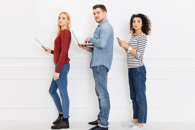 Watching you. Serious females turning heads and standing isolated on white while holding their gadgets