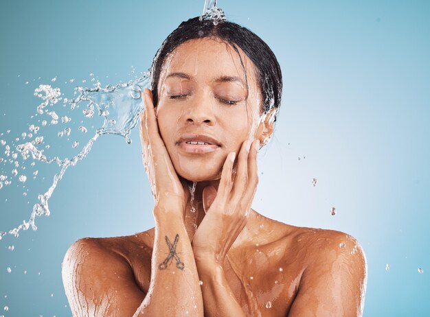 Water cleaning and shower with a black woman in studio on a blue background for hygiene or hydration Relax skincare and wellness with an attractive young female washing her skin in the bathroom