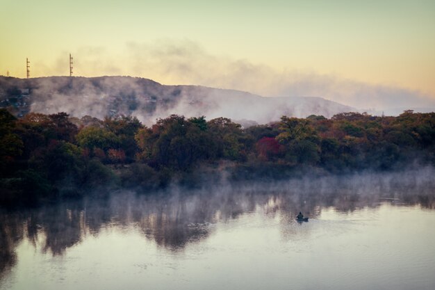 Water evapoation on the river at dawn and lonely fisher on a boat at autumn season