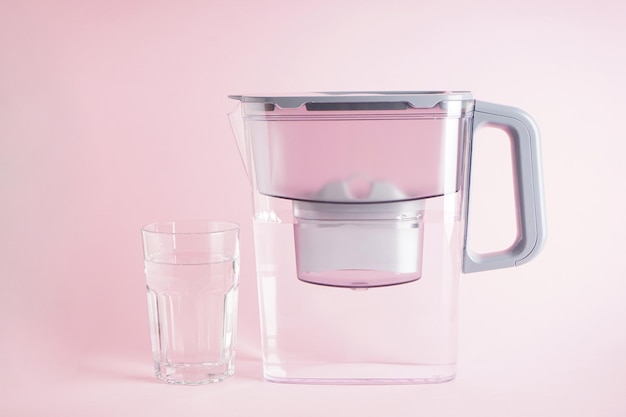 Water filter jug and a glass of water on pink background