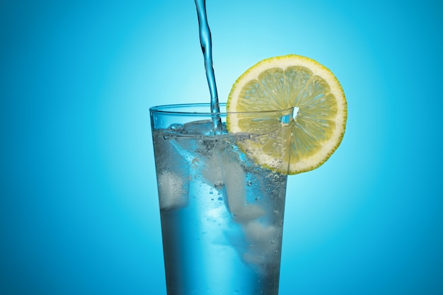 L'acqua di una bottiglia viene versata in un bicchiere con ghiaccio e limone su sfondo blu.