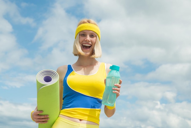 水と水分補給のコンセプトスポーツ活動屋外ブロンド陽気な笑顔の女性がスポーツヨガを保持しています...