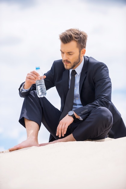 Water is leven. Depressieve jonge zakenman die fles met water vasthoudt en ernaar kijkt terwijl hij op zand zit