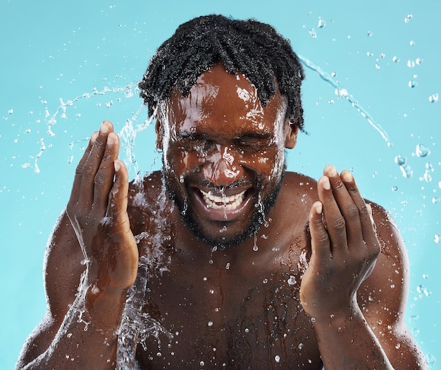 water splash cleaning and face with a model black man in studio on a blue background for hydration bathroom skincare and hygiene with a young male washing his face for natural skin treatment
