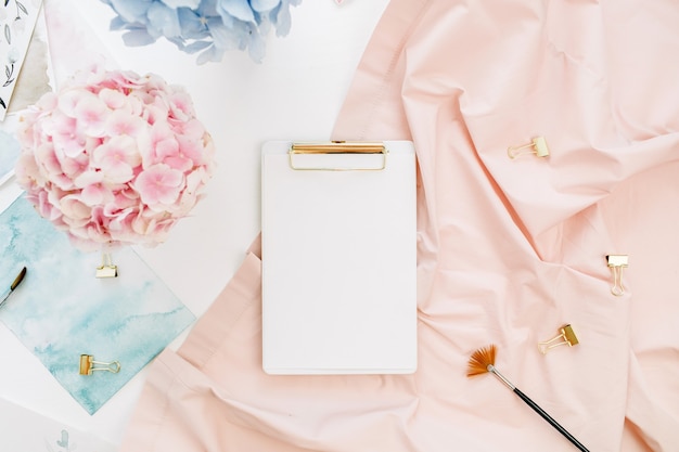 Watercolor artist home office desk workspace with copy space clipboard, pastel hydrangea flowers bouquet, watercolor painting, peachy blanket