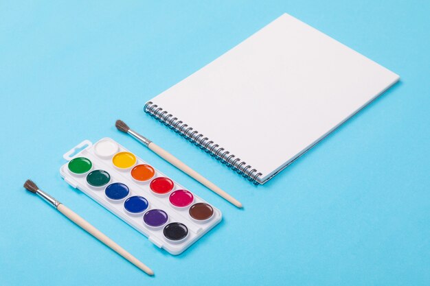 Watercolor paints and brush with album for painting isolated on blue background