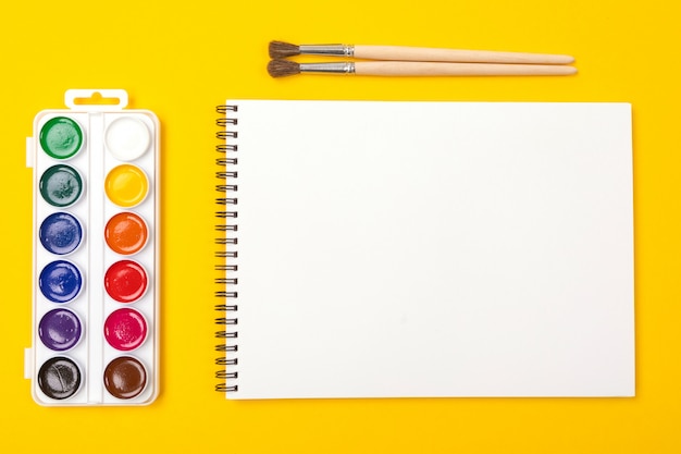 Watercolor paints and brushes with album for drawing isolated on yellow background