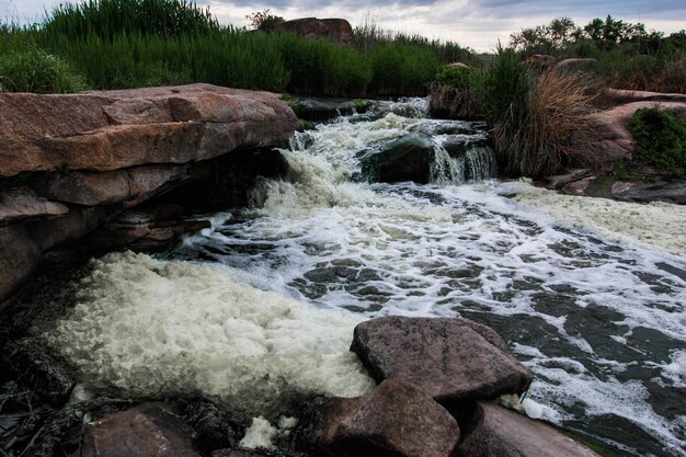 Waterfall flows among high rocks, red stones and cliffs, green bushes and trees