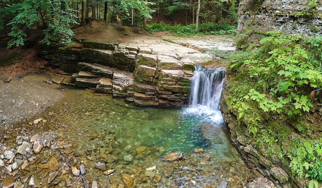 Waterfall on mountain river with white foamy water falling down from rocky formation in summer forest.