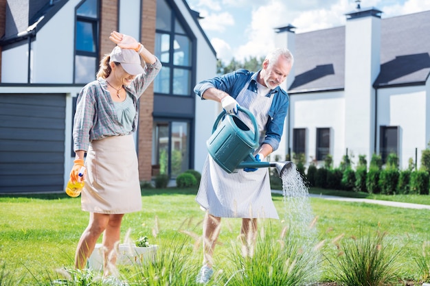 Watering bed. Husband and wife wearing casual clothes and aprons feeling lovely and happy while watering their garden bed