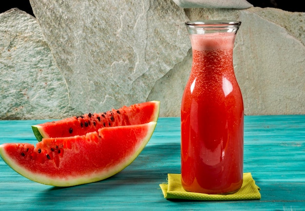 Watermelon juice in bottle and two slices of watermelon on blue wood and stone background.